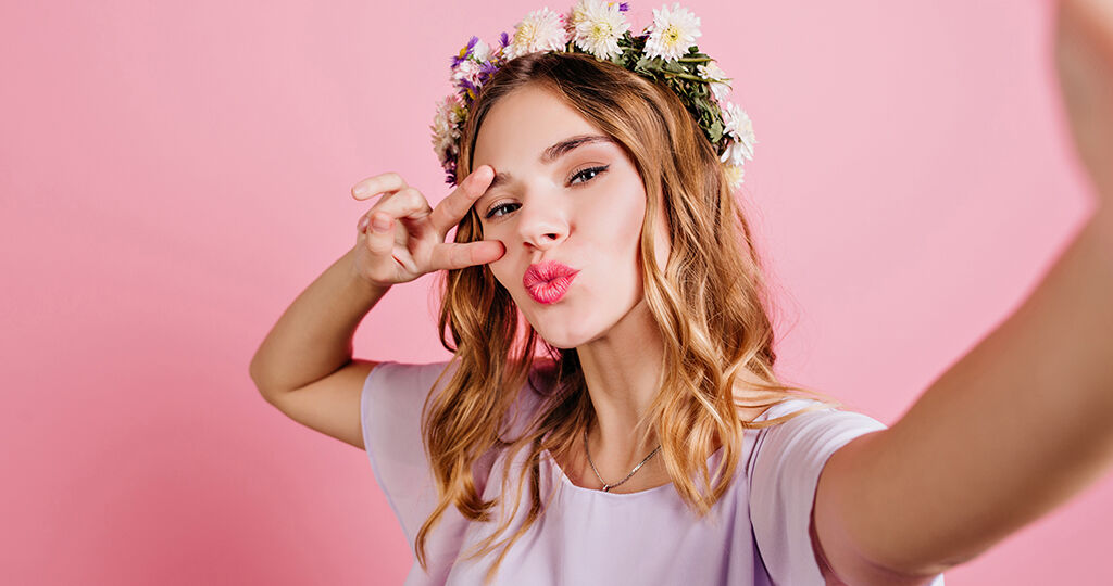 Close-up portrait of enchanting white girl wears trendy flower wreath. Charming european lady with blonde hair making selfie on pink background..